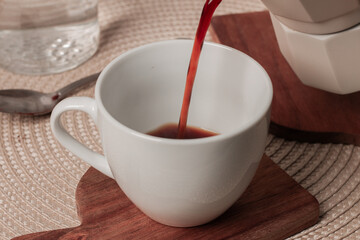 Freshly brewed coffee being poured into a white cup on a wooden coaster at a cozy breakfast table