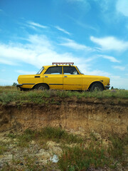 Fototapeta premium Old yellow vintage car on a hill with exposed soil layers. Retro vehicle in rural nature under a bright blue sky, nostalgic countryside atmosphere.
