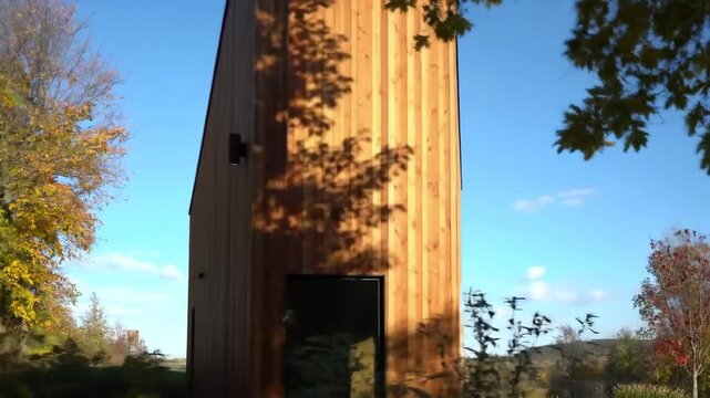 Modern wooden tower cabin stands surrounded by autumn trees under a clear blue sky.
