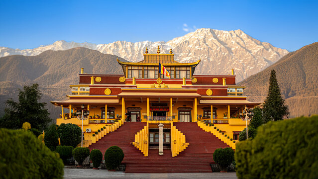 A vibrant, symmetrical view of a Tibetan Buddhist Monastery in the Dharamshala region of Himachal Pradesh, India. The architecture is striking, featuring traditional colors—a deep red base and steps