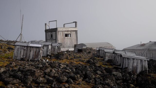 Desolate arctic outpost with weathered wooden shack and rows of abandoned dog kennels scattered across a rugged volcanic rock slope under foggy skies, amid sparse tundra