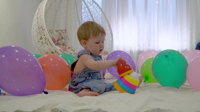 a baby in denim overalls plays with a colorful plastic pyramid sorter toy on a white bed, surrounded by festive balloons in a bright, cheerful room.