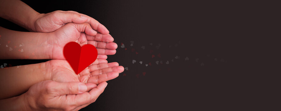 Hands of an adult and child gently holding a red heart shape together, symbolizing love, care, donation, compassion, and family support on a dark background with floating heart bokeh elements.