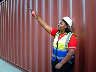 Female warehouse worker in safety vest and helmet holding up walkie-talkie for communication at container yard. Concept of logistics, teamwork, industrial safety, and diversity in workforce