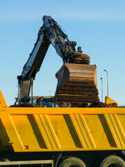 Excavator unloading soil into a yellow dump truck container at construction site on sunny day