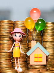 Toy girl and house with colorful balloons standing on stacks of gold coins symbolizing saving goals