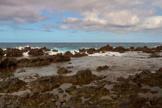 Rocky volcanic shoreline on Tenerife, where waves crash against dark lava formations under a partly cloudy sky.