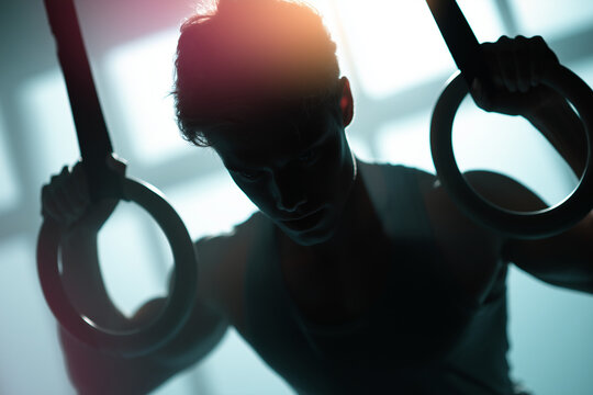 Silhouetted athlete gripping gymnastic rings in dramatic backlit gym light &mdash; intense strength training, focused determination and athletic grit