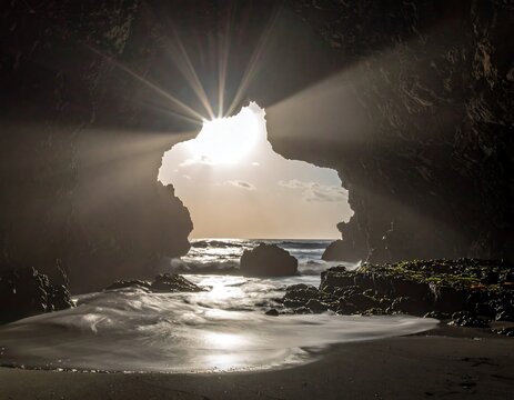 Sunlight streams through a cave opening revealing a rugged coastline. The ocean surrounds the rocky formation