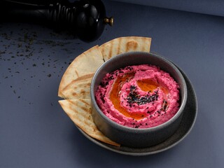 A delicious, home-made pink hummus with olive oil and black sesame seeds is served with pita bread on a modern grey table, ready to be eaten. The appetizer looks flavorful and vibrant
