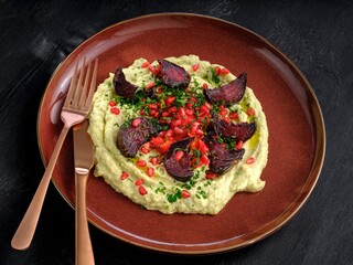 Roasted beets are artfully arranged on a bed of hummus, adorned with pomegranate seeds and fresh herbs. A rustic plate, copper cutlery, and dark background create a cozy scene