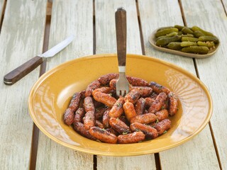 A yellow plate is filled with tiny sausages, garnished with herbs, a fork, and accompanied by a plate of pickles. A knife rests beside on a wooden surface