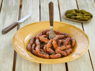 A plate of tasty looking sausages sits on a weathered table. A fork sticks out, ready to enjoy the snack. A side of pickles and a knife complete the casual outdoor meal