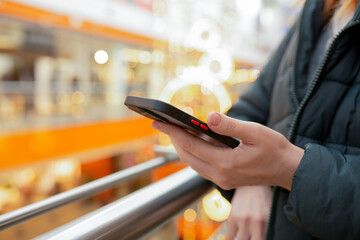 Person using a smartphone while leaning on a railing inside a shopping mall with colorful blurred lights in the background. Concept of messaging, mobile apps, navigation, technology on the go