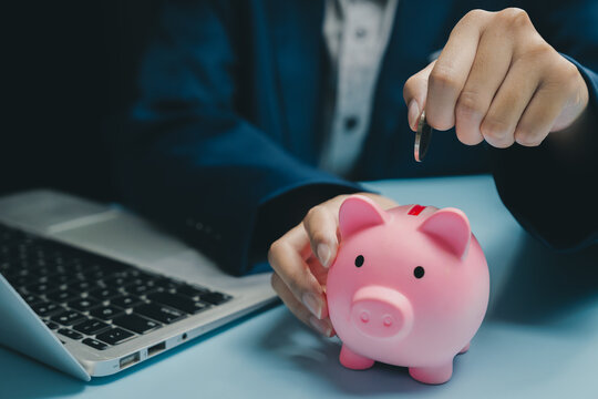 Saving money concept with person putting coin into pink piggy bank near laptop on blue table, financial planning and investment idea for future security and growth