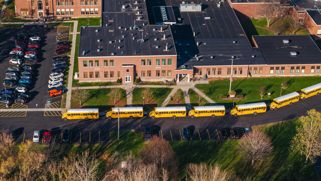 Aerial shot of an American school campus with yellow school buses lined up outside, surrounded by autumn trees and suburban greenery.