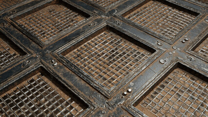 Weathered dark metal grid, featuring ornate square and diamond patterns with rusty, sandy debris underneath and decorative rivets.