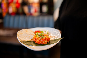 A close-up shot of a gourmet salmon tartare appetizer, meticulously plated with microgreens and bread on a white dish, held by a person in a restaurant setting