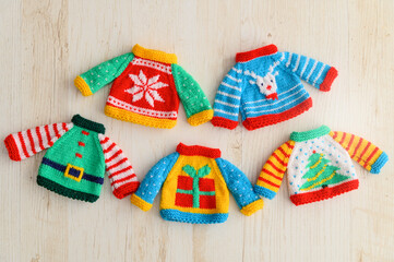 Colorful miniature knitted Christmas sweaters arranged on a light wooden surface, featuring festive patterns such as a snowflake, reindeer, gift box, Christmas tree, and an elf outfit