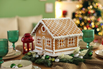 Festive table setting with beautiful gingerbread house against blurred lights indoors, closeup. Christmas atmosphere