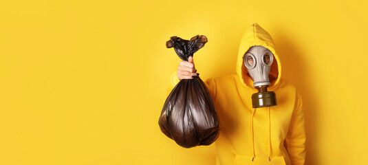 Obraz premium Individual in yellow hoodie wearing a gas mask, holding a black trash bag against a solid yellow background. Concept of pollution and waste management.