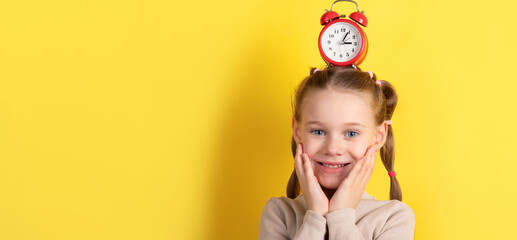 Cheerful girl with pigtails smiling and holding her face, red alarm clock balanced on head, vibrant yellow background.