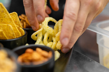 A close-up, high-angle photo of hands picking up golden onion ring snacks from a dark platter with tortilla chips and other snacks. Bright studio light