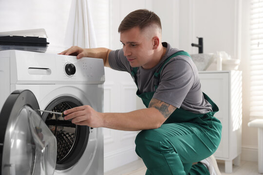 Repairman in uniform fixing broken washing machine with screwdriver at home