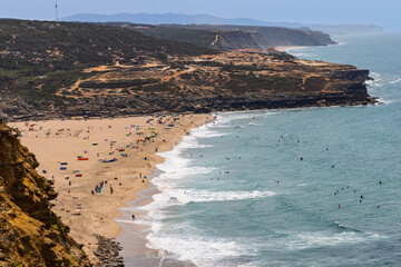 High-angle view from Miradouro da Carvoeira of Praia da Foz do Lizandro beach. People sunbathe on the sand and surf in the ocean waves.