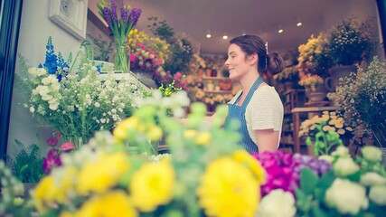 Cheerful young woman with a smile on her face is seen in her flower shop, donning an apron. The florist looks content as she carefully puts together a vibrant bouquet to showcase in the store