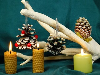Handmade Christmas decorations from pine cones with glitters, pearls and red beads hung on a white decorative tree branch, burning candles against a green backdrop 