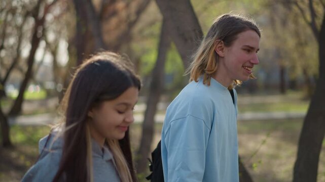 two friends walking in park exchanging awkward glances, backpacks on shoulders and autumn sunlight filtering through trees as students talk, listen and find connection in candid moment