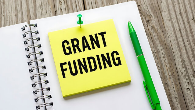 Yellow sticky note with grant funding text pinned to notebook