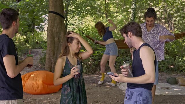 A group of young friends enjoys a relaxing day outdoors, sharing drinks and laughter. They gather closely to capture a happy moment with a selfie, celebrating their friendship amidst lush greenery.