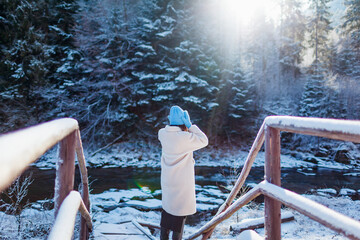 Back view of woman admiring snowy forest and river view wearing white warm fur coat. Winter landscape. Traveling for holidays