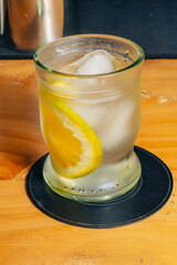Glass of lemon water with ice on black coaster over wooden surface.