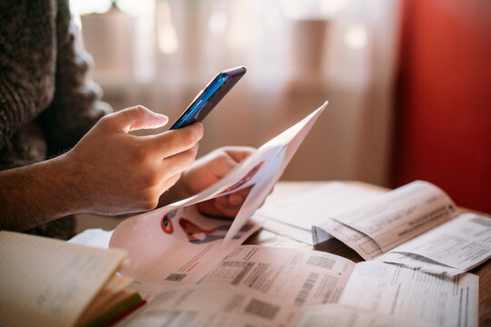 A man pays utility bills online using a QR code in an app using a smartphone in the kitchen. Close-up of hands, phone, and bills.