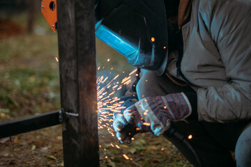 A young man is working as a gas welder on the street. A man wearing a protective mask is welding iron rungs to pillars. The worker, holding a welding machine, sparks fly around him.