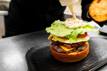 A chef adds sauce to a gourmet cheeseburger with a fried egg, cheese, and fresh lettuce, preparing a delicious meal in a commercial kitchen setting