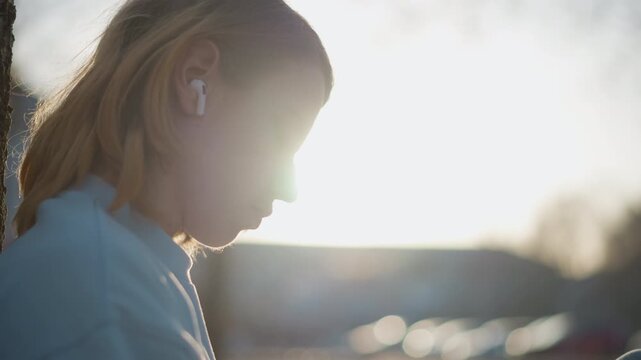 white teen leaning against tree listening to music, student taking reflective break during golden sunset, earbuds in ears, soft backlit profile, calm mood, nature background, backpack at feet