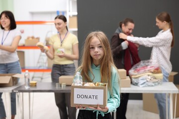 Volunteers helping homeless people in shelter, focus on girl with box of donations