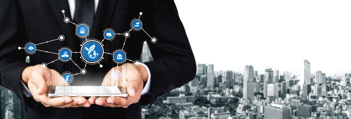 A businessman in a formal suit holds a tablet displaying digital icons related to technology and networking over a vibrant cityscape, symbolizing innovation and progress. Tessel