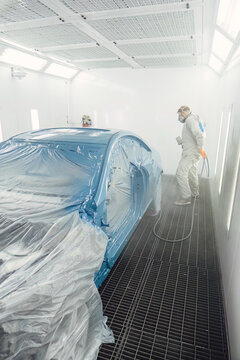 Professional painter in a protective suit spraying paint onto the car body inside a clean, modern spray booth. High quality automotive repair and bodywork service maintenance for vehicles.