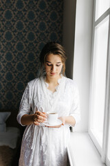 The bride in a white robe stands by the window holding a cup of coffee. She smiles softly, enjoying a calm moment before the wedding. A warm and peaceful morning atmosphere in a cozy room.
