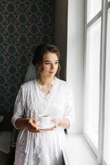 The bride in a white robe stands by the window holding a cup of coffee. She smiles softly, enjoying a calm moment before the wedding. A warm and peaceful morning atmosphere in a cozy room.