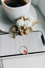 Wedding details on a white table: a cup of coffee, a smartphone with a marked date, a ring, and a small floral boutonniere. A calm and meaningful moment of preparation for the special day.