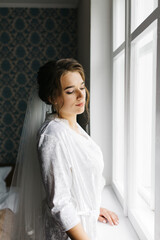 The bride in a white robe stands by the window holding a cup of coffee. She smiles softly, enjoying a calm moment before the wedding. A warm and peaceful morning atmosphere in a cozy room.