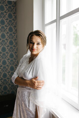 The bride in a white robe stands by the window holding a cup of coffee. She smiles softly, enjoying a calm moment before the wedding. A warm and peaceful morning atmosphere in a cozy room.