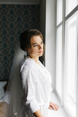 The bride in a white robe stands by the window holding a cup of coffee. She smiles softly, enjoying a calm moment before the wedding. A warm and peaceful morning atmosphere in a cozy room.