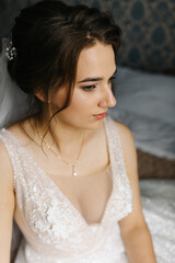 Portrait of a bride sitting indoors in her wedding dress. She looks at the camera with a calm, confident expression. Soft light highlights the lace details, veil, and floral bracelet, creating 